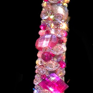 May include: A pink and clear crystal headband with a variety of shapes and sizes of crystals. The headband is made of a flexible metal band.