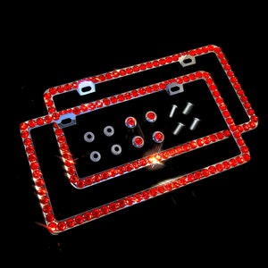May include: Two license plate frames with a chrome finish, each encrusted with vibrant red rhinestones. The frames are accompanied by mounting hardware, including screws and washers, against a black background. A decorative car accessory.
