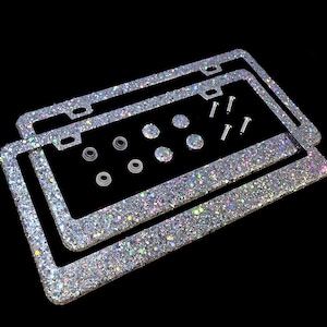 May include: Two glitter-covered license plate frames with mounting hardware. The frames are silver with iridescent glitter, and the hardware includes screws, washers, and decorative gems. The background is black.