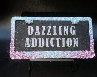 May include: A license plate frame with the words "DAZZLING ADDICTION" in silver glitter lettering. The frame is black with a border of light blue glitter and pink rhinestones.