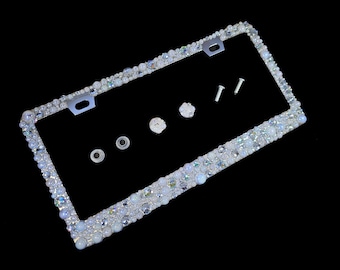 May include: A silver license plate frame with a border of white and iridescent rhinestones. The frame is designed to hold a standard license plate and includes two screws for installation.