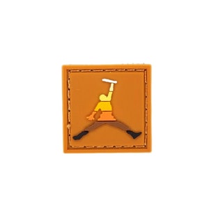 Puede incluir: Un parche cuadrado naranja con una figura estilizada en pleno salto, sosteniendo un objeto sobre su cabeza. La figura es amarilla, naranja y marrón, con zapatos negros. El parche tiene un borde cosido.