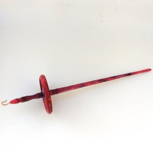 K&ouml;nnte beinhalten: Eine Handspindel aus Holz zum Spinnen von Garn. Die Spindel hat einen rot gef&auml;rbten Schaft und einen kreisf&ouml;rmigen Wirtel. Der Schaft hat einen Haken am Ende. Die Spindel ist etwa 30 cm lang.