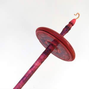 K&ouml;nnte beinhalten: Eine Handspindel mit einem roten und violetten Holzschaft und Wirtel. Der Wirtel hat ein kreisf&ouml;rmiges Design. Die Oberseite der Spindel hat einen Kupferhaken und eine rote Spitze. Die Spindel wird zum Handspinnen von Garn verwendet.