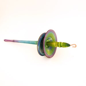 May include: A hand-painted wooden spindle in shades of green, blue, and purple. The spindle has a long, slender shaft and two large, circular whorls. A copper hook is attached to the end. This tool is used for spinning yarn.