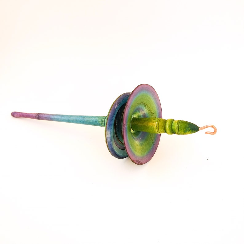 May include: A hand-painted wooden spindle in shades of green, blue, and purple. The spindle has a long, slender shaft and two large, circular whorls. A copper hook is attached to the end. This tool is used for spinning yarn.
