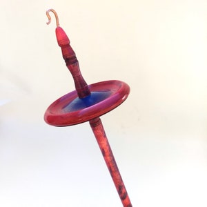 K&ouml;nnte beinhalten: Eine Handspindel mit Kupferhaken, einem rot gebeizten Holzschaft und einem kreisf&ouml;rmigen Wirtel mit roten und blauen Akzenten. Die Spindel ist zum Handspinnen von Garn und Faser bestimmt.