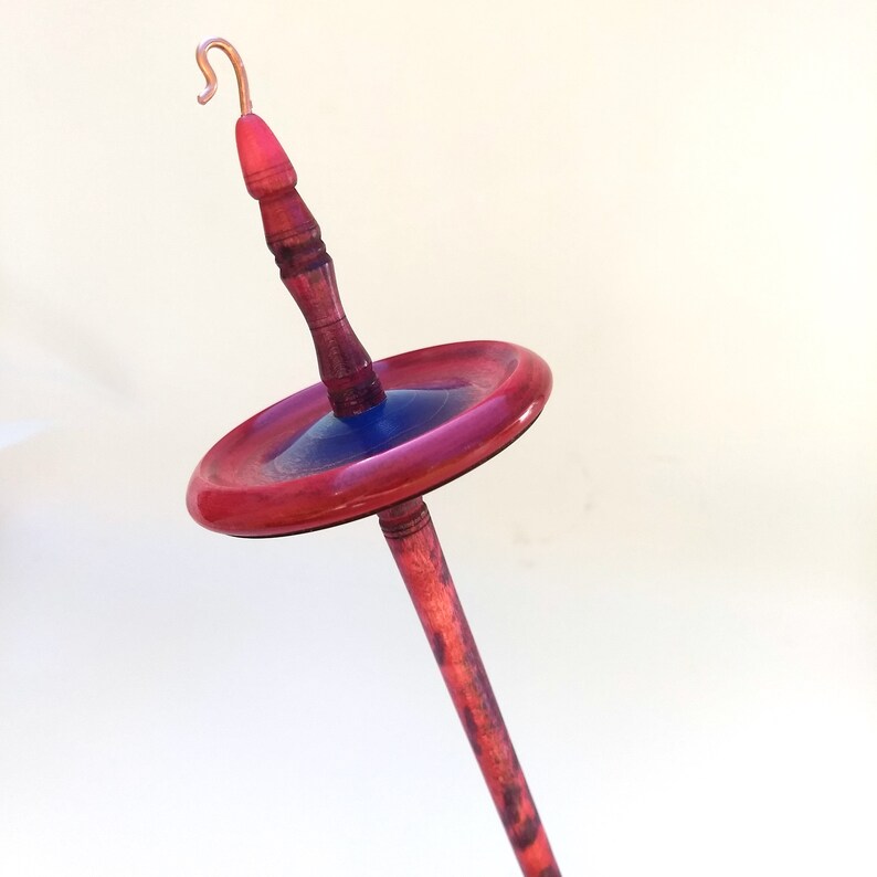 K&ouml;nnte beinhalten: Eine Handspindel mit Kupferhaken, einem rot gebeizten Holzschaft und einem kreisf&ouml;rmigen Wirtel mit roten und blauen Akzenten. Die Spindel ist zum Handspinnen von Garn und Faser bestimmt.
