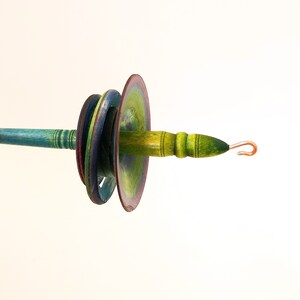 May include: A hand-painted wooden spindle with a copper hook. The spindle features a series of colorful discs in shades of green, blue, and purple. The shaft is painted in shades of green and yellow.