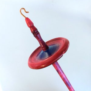 K&ouml;nnte beinhalten: Eine Handspindel mit einem roten und violetten Holzschaft und Wirtel. Der Wirtel hat einen roten Au&szlig;enring und eine blaue Mitte. Die Oberseite der Spindel hat einen Kupferhaken. Dieses Werkzeug wird zum Handspinnen von Garn verwendet.