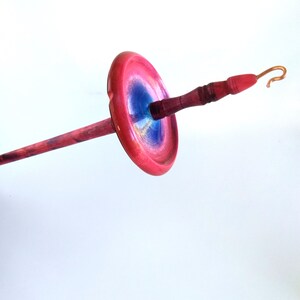 K&ouml;nnte beinhalten: Eine Handspindel mit einem rot-blauen, scheibenf&ouml;rmigen Wirtel und einem Holzschaft. Der Wirtel hat eine leuchtend rote Au&szlig;enseite mit einem blauen Zentrum. Schaft und Griff sind tiefrotbraun, der Haken kupferfarben.