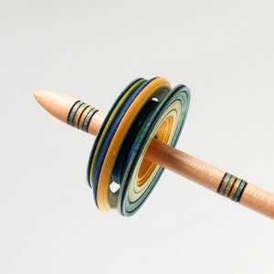May include: A wooden drop spindle with a light brown shaft and a colourful whorl. The whorl features layers of blue, yellow, and green, creating a vibrant, layered effect. The spindle is designed for spinning yarn.