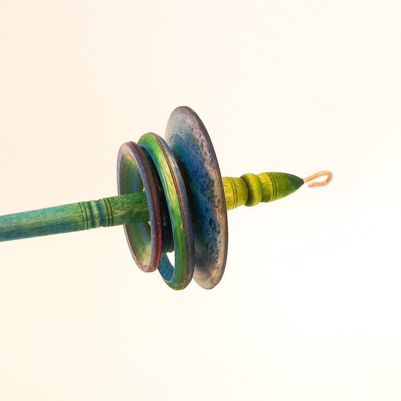 May include: A hand spindle with a teal shaft and a series of colorful, graduated discs. The discs transition from red to green to blue, with a copper hook at the end. The wooden spindle is a tool for hand spinning yarn.