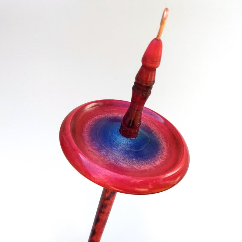 K&ouml;nnte beinhalten: Eine Handspindel zum Spinnen von Garn. Die Holzspindel hat einen lebendigen Farbverlauf, der von Rot nach Blau &uuml;bergeht. Die Spindel hat einen runden, flachen Wirtel und einen schlanken Schaft. Die Gesamth&ouml;he betr&auml;gt ca. 20 cm.