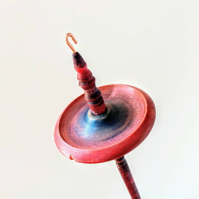 K&ouml;nnte beinhalten: Eine Handspindel, ein Werkzeug zum Spinnen von Garn, mit einem roten und schwarzen Holzschaft und Wirtel. Der Wirtel hat eine rot, blau und violett schillernde Oberfl&auml;che. Oben an der Spindel befindet sich ein Kupferhaken.