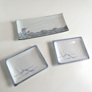 Puede incluir: Tres platos rectangulares de cerámica con superficies blancas y detalles pintados en azul. Un plato grande presenta un velero sobre olas estilizadas, mientras que dos platos más pequeños muestran delicados dibujos azules de juncos.