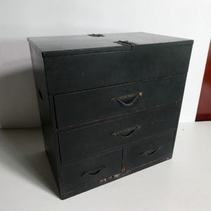 May include: A dark charcoal gray wooden chest featuring a hinged top lid and four drawers. The two upper drawers are full-width, while the two lower drawers are side-by-side. Each drawer is equipped with a simple, curved metal pull handle.