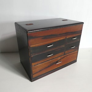 Könnte beinhalten: Eine Holztruhe mit einer dunkelbraunen und orangefarbenen Holzmaserung, die fünf Schubladen unterschiedlicher Größe aufweist. Jede Schublade ist mit einem kleinen, horizontalen silberfarbenen Metallgriff ausgestattet. Die Oberseite ist flach, dunkelbraun und hat zwei Scharniere.