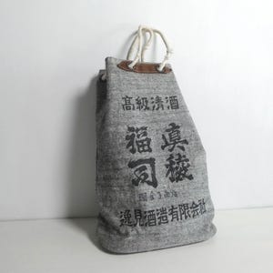 Op de afbeelding: Een grijze tas van gestructureerde stof met een taps toelopende bovenkant en witte touwgrepen bevestigd aan bruine leren lipjes. De tas is aan de voorzijde bedrukt met zwarte Japanse kanji-tekens, waaronder de tekens voor hoogwaardige sake.