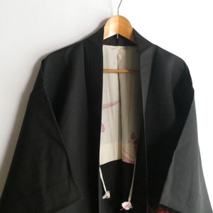 Puede incluir: Un kimono japonés negro con un forro floral blanco y rosa. El kimono está colgado en una percha de madera.