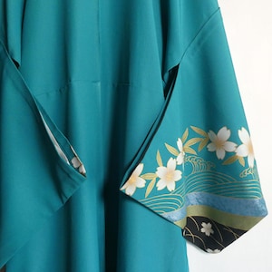 May include: A teal kimono featuring wide sleeves with a decorative pattern of white cherry blossoms, gold leaves, and stylized blue and black waves along the lower edges. The garment has a smooth, solid-colored teal fabric throughout the main body.