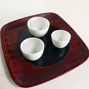 May include: Three small, white ceramic bowls sit on a black circular surface, set within a square, red-brown tray. The tray has a textured, woven-like pattern around the edges.