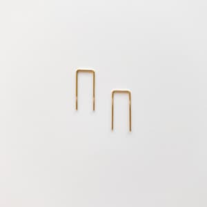 May include: Two gold-colored, minimalist U-shaped earrings on a white background. The earrings are simple, geometric, and modern in design. The earrings are small and delicate, with a polished finish. The earrings are likely made of metal.