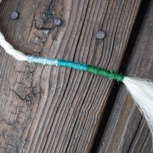 Puede incluir: Una extensión de cabello blanca con una sección trenzada verde, azul verdoso y azul.