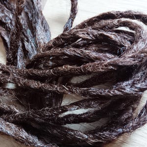 Puede incluir: Un manojo de extensiones de cabello sintético de color marrón oscuro con una textura retorcida similar a una cuerda. Los mechones individuales están estrechamente entrelazados para formar fibras largas y flexibles de color marrón oscuro.