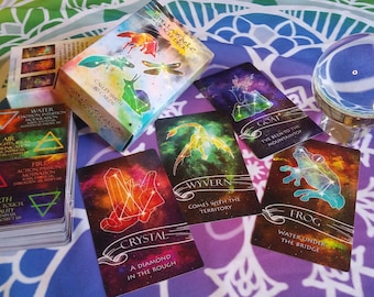 May include: A set of oracle cards with a celestial theme. The cards feature colorful illustrations of constellations, crystals, and mythical creatures. The box for the cards has a watercolor design with a dragonfly, a snail, and a bird.