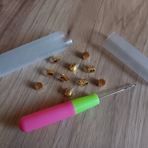 Może przedstawiać: Zestaw złotych metalowych koralik&oacute;w, r&oacute;żowo-zielony szydełko i dwa przezroczyste plastikowe pojemniki do przechowywania.