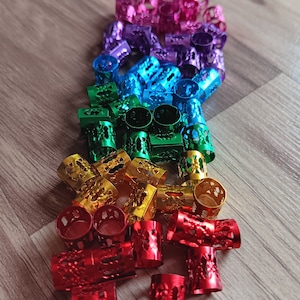 May include: A rainbow assortment of colorful metal hair beads. The beads are arranged in a rainbow pattern, starting with pink and ending with red. The beads are all the same size and shape, and they have a decorative design.