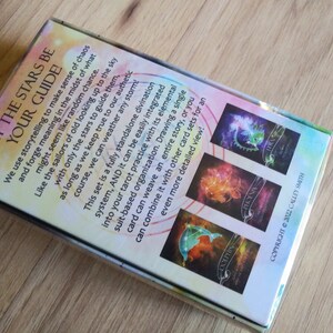 May include: A box of Let the Stars Be Your Guide oracle cards. The box features a colorful design with a starry sky and constellations. The back of the box includes a description of the cards and how to use them.