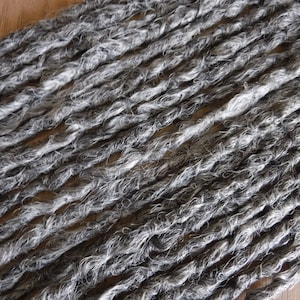 May include: A close-up of a bundle of gray and white synthetic dreadlocks. The dreadlocks are made of a soft, textured material and are arranged in a neat, even row.