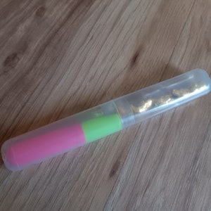 Może przedstawiać: Przezroczysty plastikowy tubus z r&oacute;żowym i zielonym markerem w środku. Tubus jest otwarty z jednej strony i zawiera złotą folię.