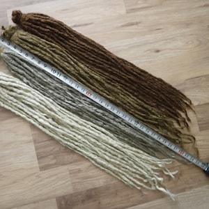May include: Three strands of synthetic dreadlocks in different colours: light brown, dark brown, and white. The dreadlocks are laid out on a wooden surface with a measuring tape running along the length of the strands. The tape measures up to 86 cm.