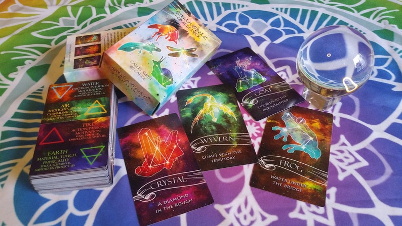 May include: A set of oracle cards with a celestial theme. The cards feature colorful illustrations of constellations, crystals, and mythical creatures. The box for the cards has a watercolor design with a dragonfly, a snail, and a bird.