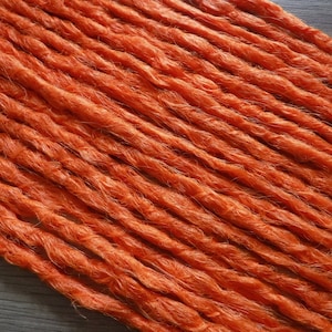 May include: Close-up of several orange dreadlocks, arranged diagonally across the frame. The synthetic hair appears textured and slightly coarse, with a vibrant, solid color. The dreadlocks are likely for hair extensions or styling.