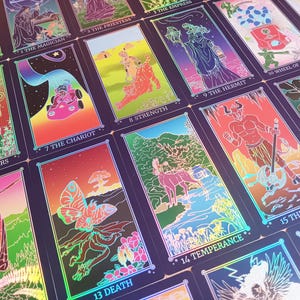 May include: A collection of tarot cards with vibrant illustrations and holographic accents. Each card features unique artwork and titles such as "The Lovers," "The Chariot," and "The Devil." The cards are arranged in a visually appealing display.