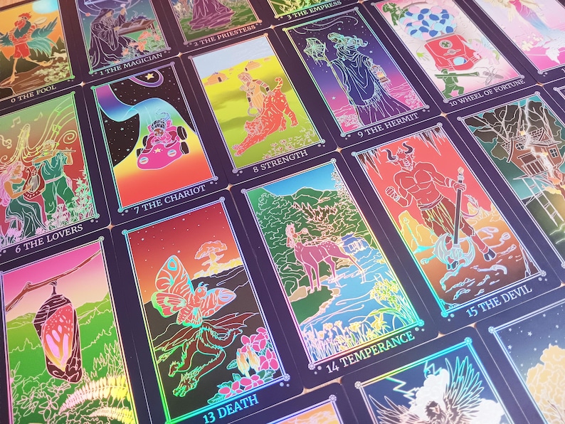 May include: A collection of tarot cards with vibrant illustrations and holographic accents. Each card features unique artwork and titles such as "The Lovers," "The Chariot," and "The Devil." The cards are arranged in a visually appealing display.