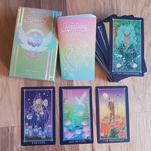 May include: A Heartsong Tarot deck with a box and guidebook. The box is green and gold with the text "Pandemonia Heartsong Tarot". Three tarot cards are visible, including "9 of Cups", "Ace of Cups", and "7 of Pentacles". The deck is on a wooden surface.