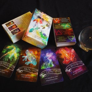 May include: A set of Stella Maris Oracle Cards with a crystal ball and a guidebook. The cards feature colorful illustrations of constellations and mythical creatures, such as a wyvern, frog, goat, and crystal. The guidebook has a title that reads "Stella Maris Oracle Deck" and "Cally Smith Cards".