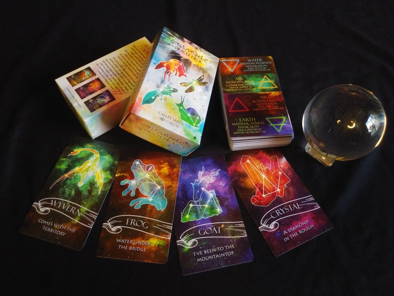May include: A set of Stella Maris Oracle Cards with a crystal ball and a guidebook. The cards feature colorful illustrations of constellations and mythical creatures, such as a wyvern, frog, goat, and crystal. The guidebook has a title that reads "Stella Maris Oracle Deck" and "Cally Smith Cards".