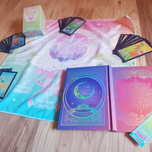 May include: A collection of tarot cards, books, and a colorful cloth with celestial and geometric designs. The set includes a deck of cards, two books with intricate covers, and a rectangular box. The color palette includes blues, pinks, and greens.
