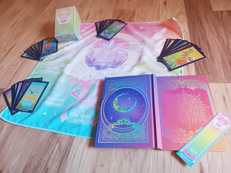 May include: A collection of tarot cards, books, and a colorful cloth with celestial and geometric designs. The set includes a deck of cards, two books with intricate covers, and a rectangular box. The color palette includes blues, pinks, and greens.