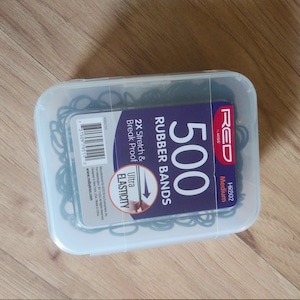Może przedstawiać: Przezroczysty plastikowy pojemnik wypełniony 500 czarnymi gumkami. Pojemnik ma etykietę z napisem "RED" i "500 Rubber Bands" z tekstem "2X Stretch & Break Proof" i "Ultra Elasticity".
