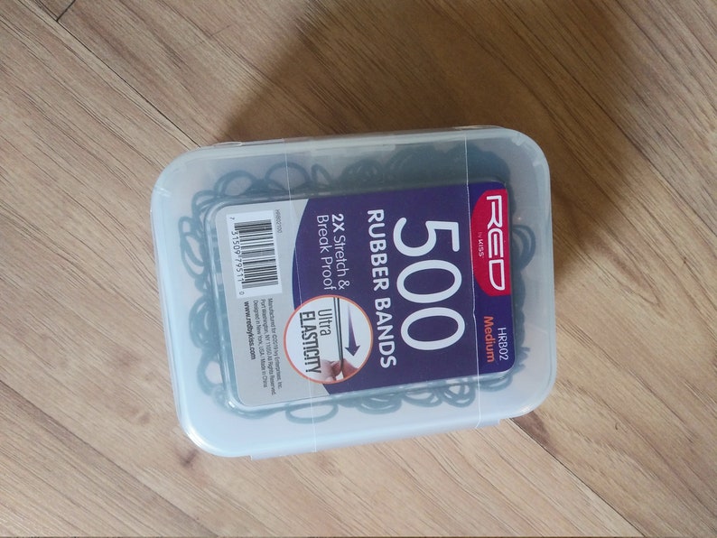 Może przedstawiać: Przezroczysty plastikowy pojemnik wypełniony 500 czarnymi gumkami. Pojemnik ma etykietę z napisem "RED" i "500 Rubber Bands" z tekstem "2X Stretch & Break Proof" i "Ultra Elasticity".