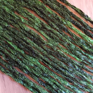 Könnte beinhalten: Nahaufnahme von mehreren grünen und schwarzen Dreadlocks, die diagonal auf einer hellbraunen Oberfläche angeordnet sind. Die Dreadlocks haben ein strukturiertes, natürliches Aussehen mit verschiedenen Grün- und Schwarztönen.