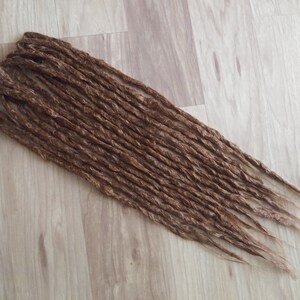 Peut inclure: Un ensemble de dreadlocks bruns, disposés sur une surface claire imitation bois. Les dreadlocks sont d'une couleur marron moyen et semblent faits à la main. La texture des dreadlocks est visible, avec des mèches individuelles créant un aspect naturel.