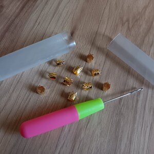 Może przedstawiać: Zestaw złotych koralik&oacute;w do włos&oacute;w w dw&oacute;ch przezroczystych plastikowych pojemnikach, wraz z r&oacute;żowym i zielonym haczykiem do szydełka.